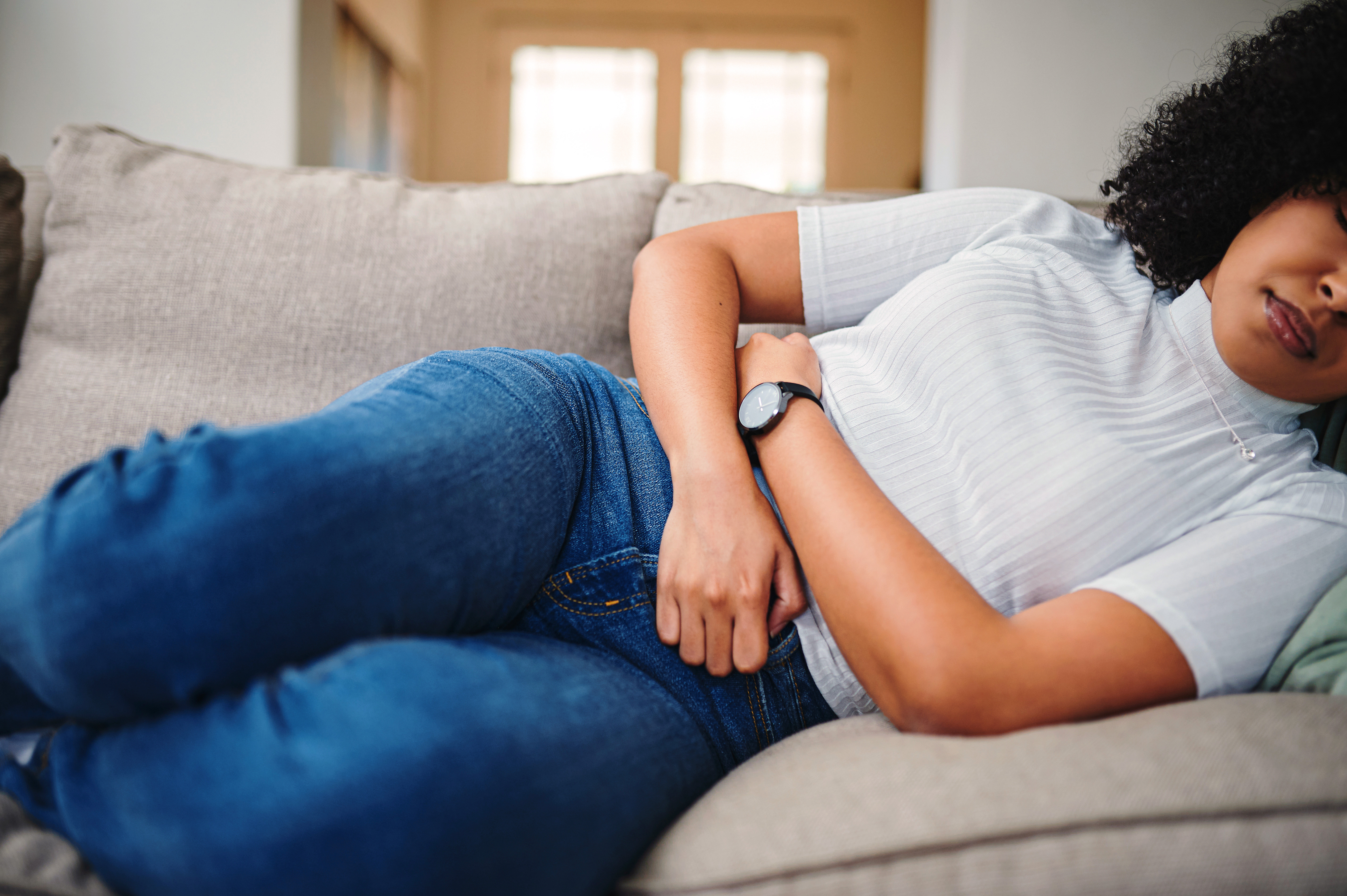 A woman holding her stomach in discomfort while laying on the couch at home.