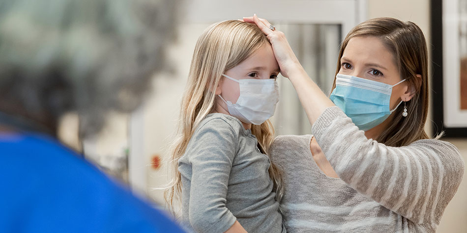 A mother speaks to a pediatrician, while carrying her young daughter. She rests her hand on her daughter's forehead to check her fever. Both are wearing face masks.