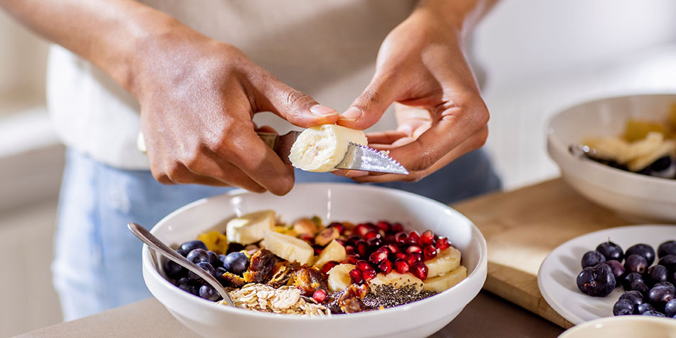 A person cuts a banana with a knife into a mixed fruit and oats bowl.