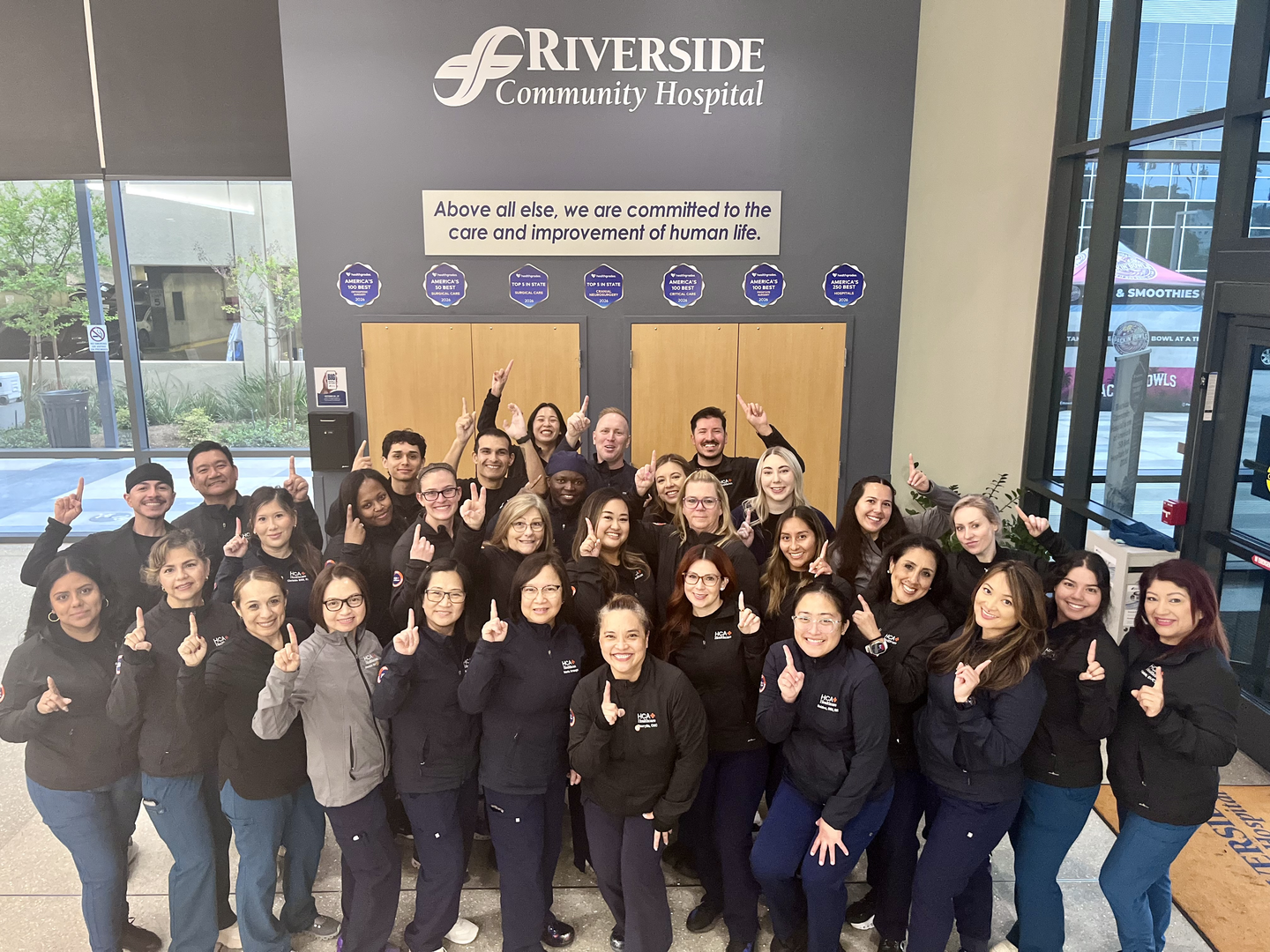 Group photo of Riverside Community Hospital care team members standing in the hospital lobby beneath the hospital name and mission statement, smiling and holding up one finger to celebrate G1 Advanced Surgical Telemetry Unit being named a 2025 HCA Healthcare Unit of Distinction Top Ranking Unit for medical-surgical care.