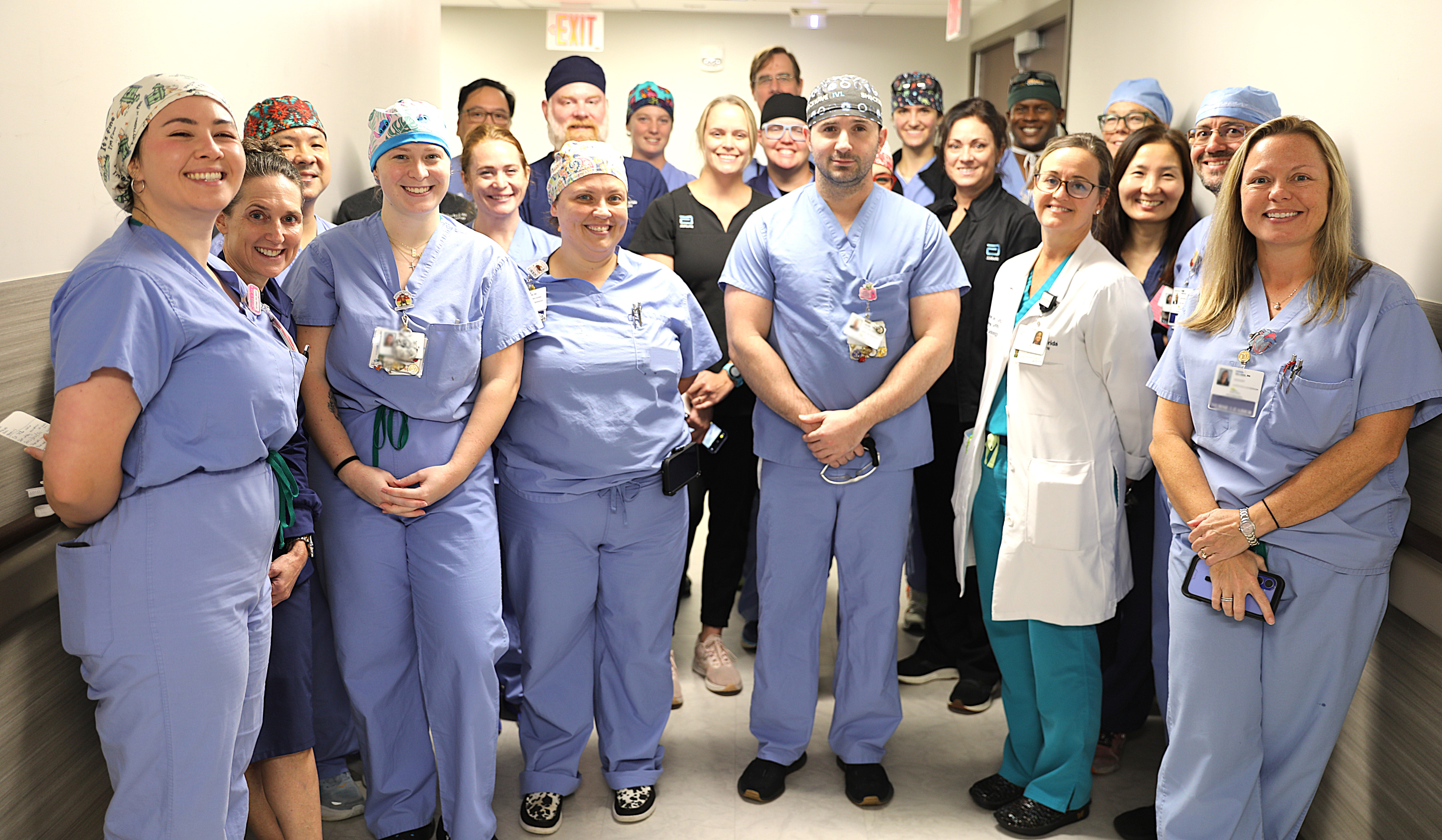 23 people, mostly women, dressed in light blue hospital scrubs standing in a huddle in a hallway.