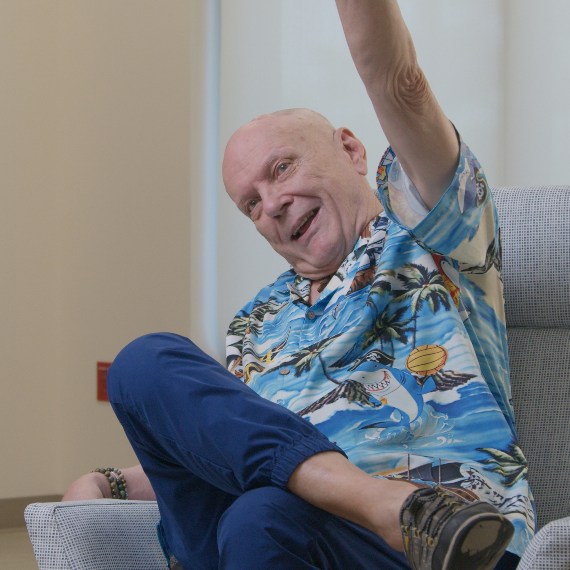 Larry Mundt in a bright blue Hawaiian shirt, sitting in a chair raising his left hand to the sky.