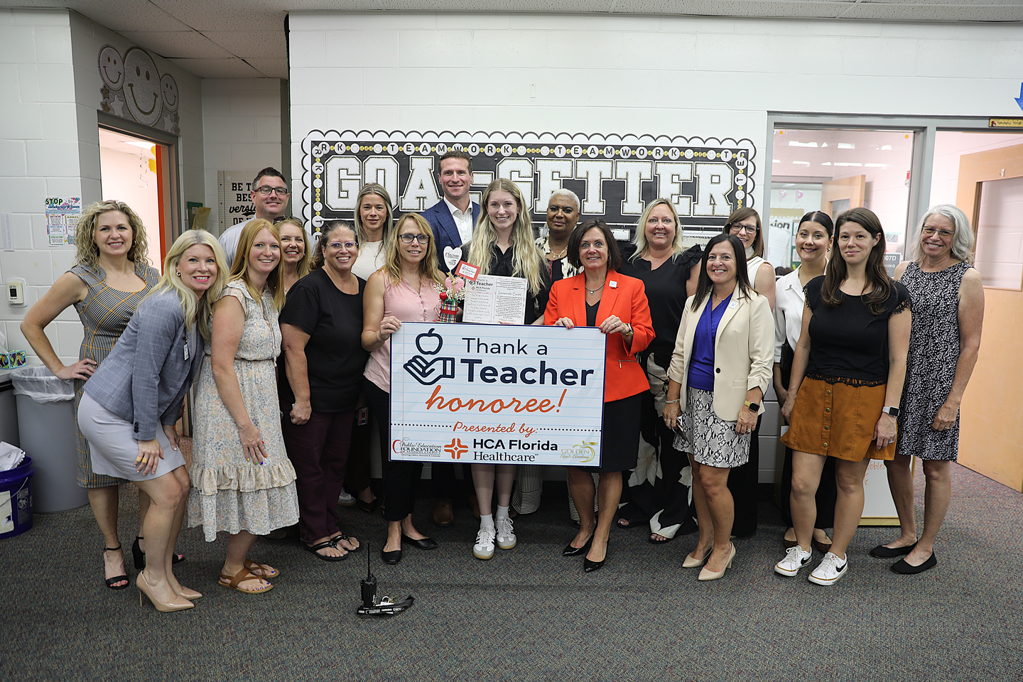 A group of approximately 18 educators, school staff, and healthcare representatives stand together inside an elementary school classroom, celebrating a "Thank a Teacher" honoree. At the center, a young teacher holds a certificate and a handmade award while several attendees display a large sign reading "Thank a Teacher honoree!" presented by HCA Florida Healthcare. A classroom bulletin board with the word "GOAL-GETTER" is visible in the background.