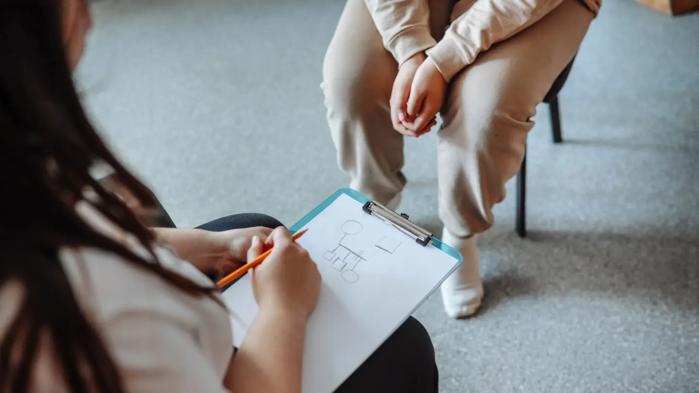 Psychologist's hands draw genogram on clipboard. Teen patient sits opposite with folded arms.