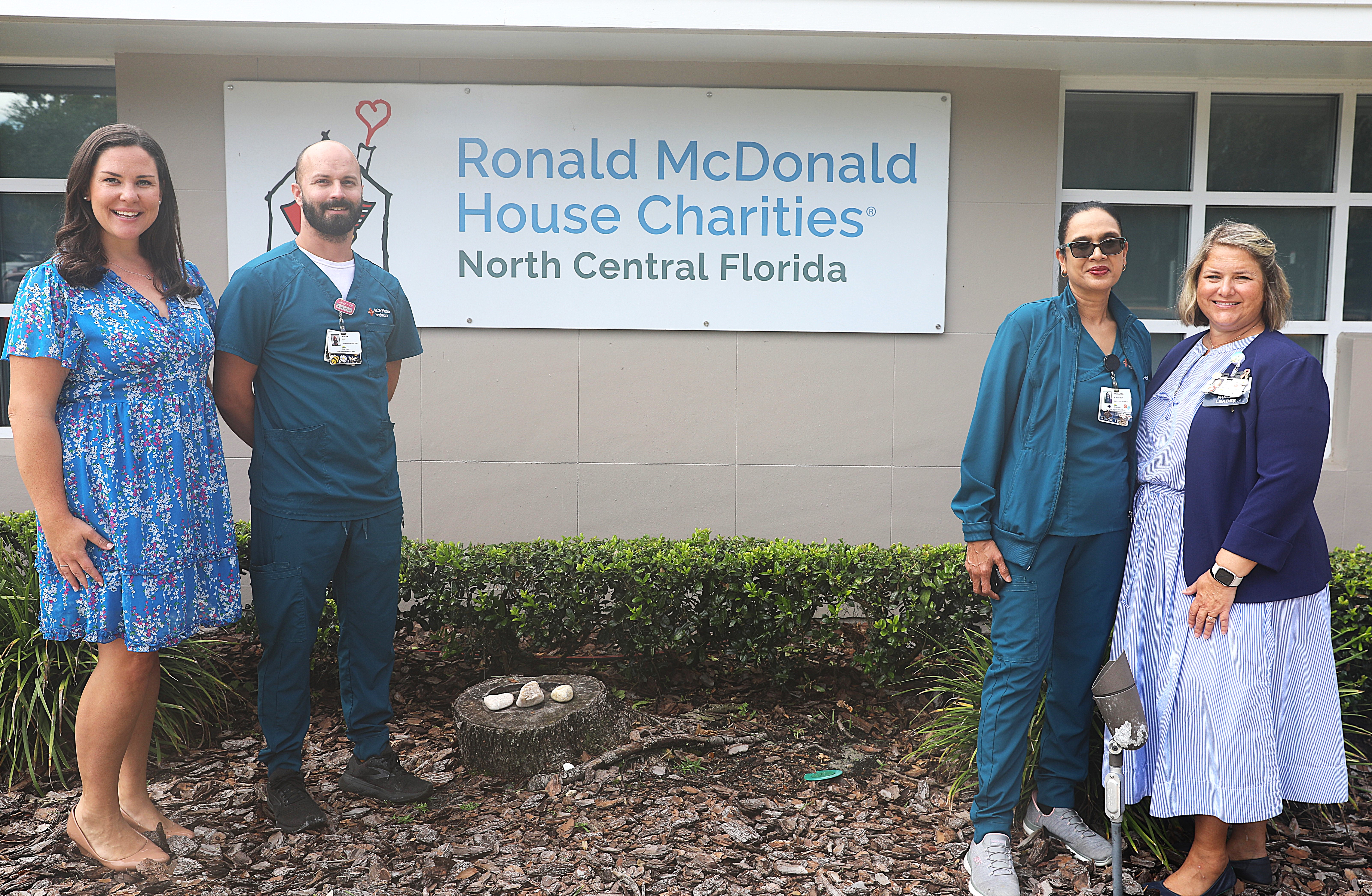 A group of three women and one man are smiling and standing in a line in front of a sign that reads Ronald McDonald House Charities of North Central Florida.