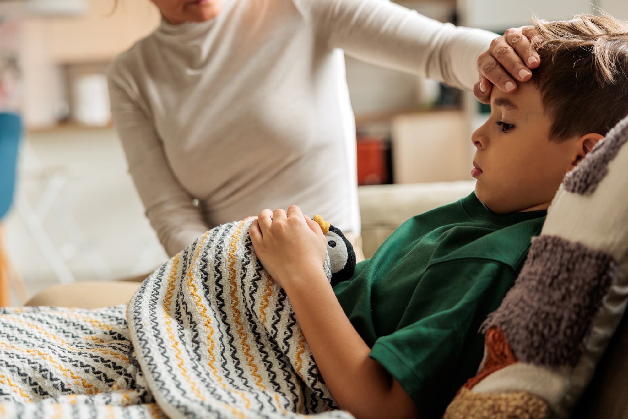 Adult caregiver checking a young boy’s forehead for fever as he lies on the couch under a blanket holding a stuffed penguin.