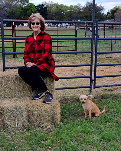 Jacquelyn Jones wearing sunglasses and sitting on a hay bale outside a gate, with her dog nearby.