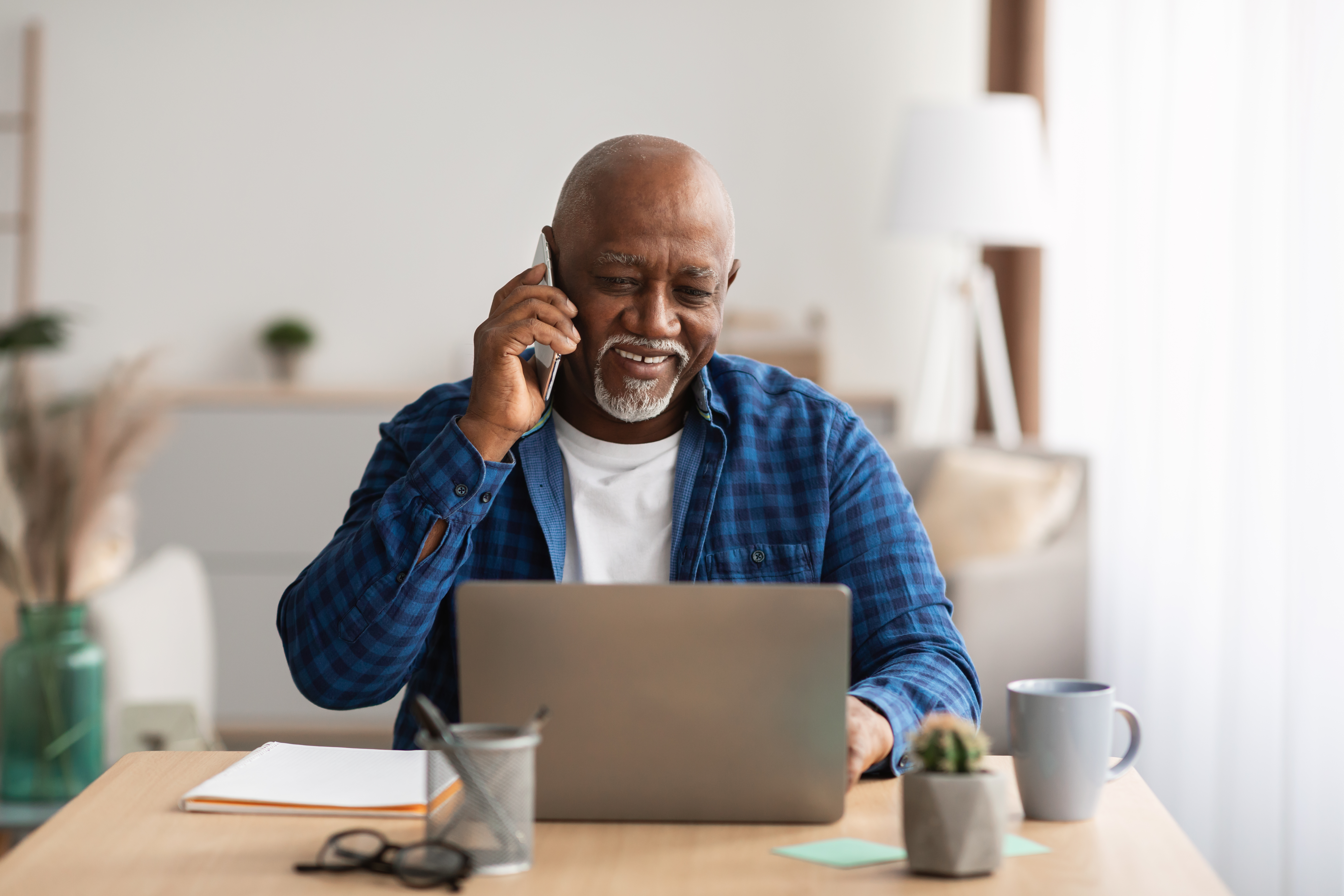 A senior male sitting in front of a laptop, smiling and talking on a phone.