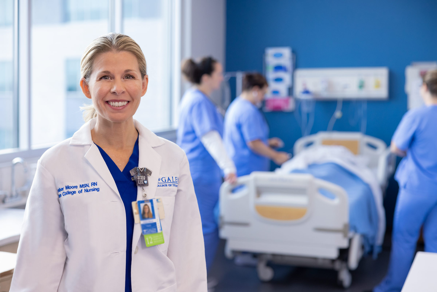 A smiling nursing instructor wearing a white lab coat and ID badge stands in the foreground of a clinical simulation lab. Behind her, several students in blue scrubs work around a hospital bed in a patient-care training environment. Large windows provide natural light, and medical equipment is visible against a blue accent wall.