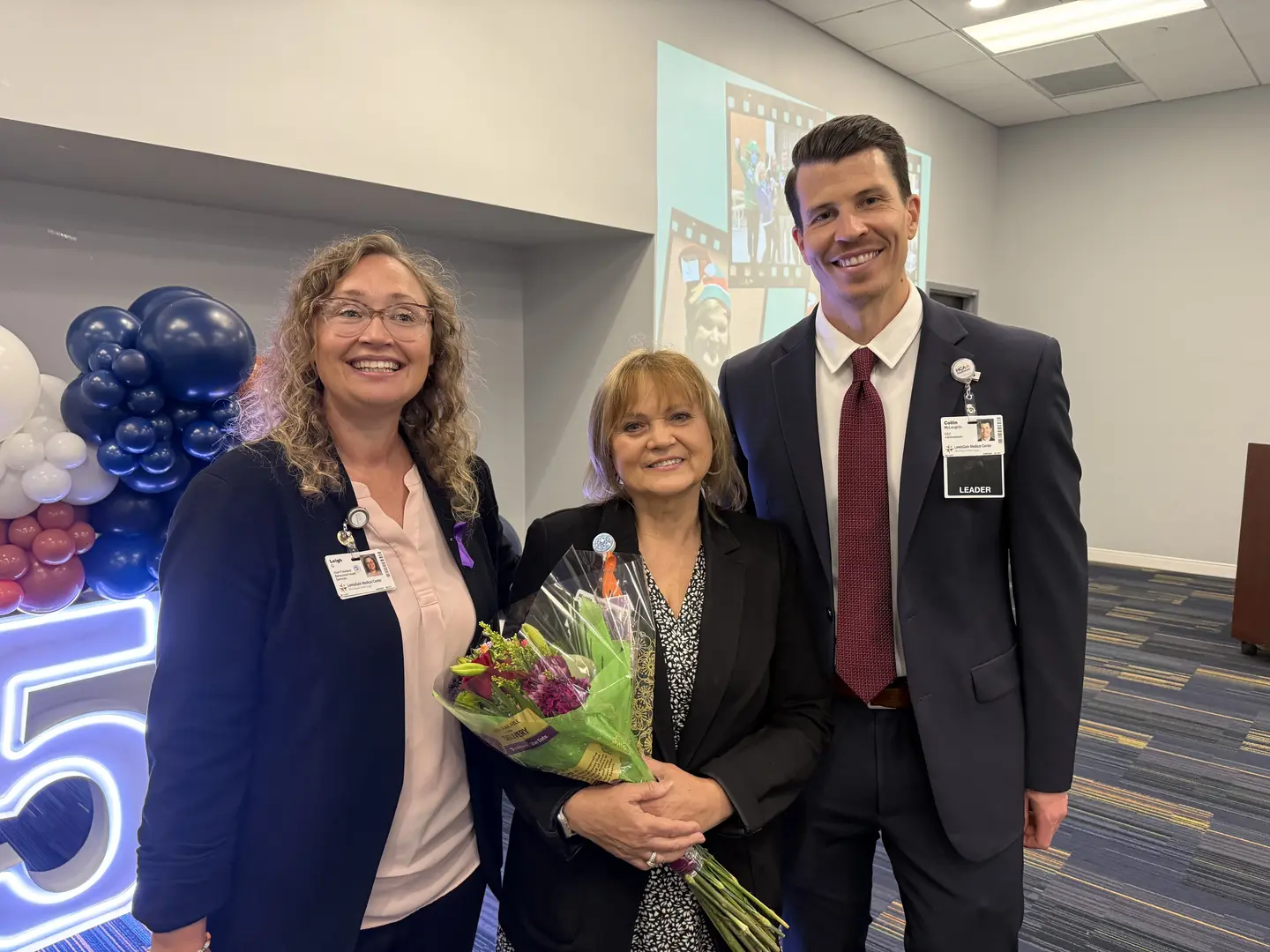 Three people stand side-by-side looking at the camera. They are each smiling. A woman wearing a suit is on the left. A woman wearing a dark colored jacket and holding flowers is in the middle. A man wearing a dark suit and tie is standing to the right of the woman holding the flowers. There are balloons behind them.