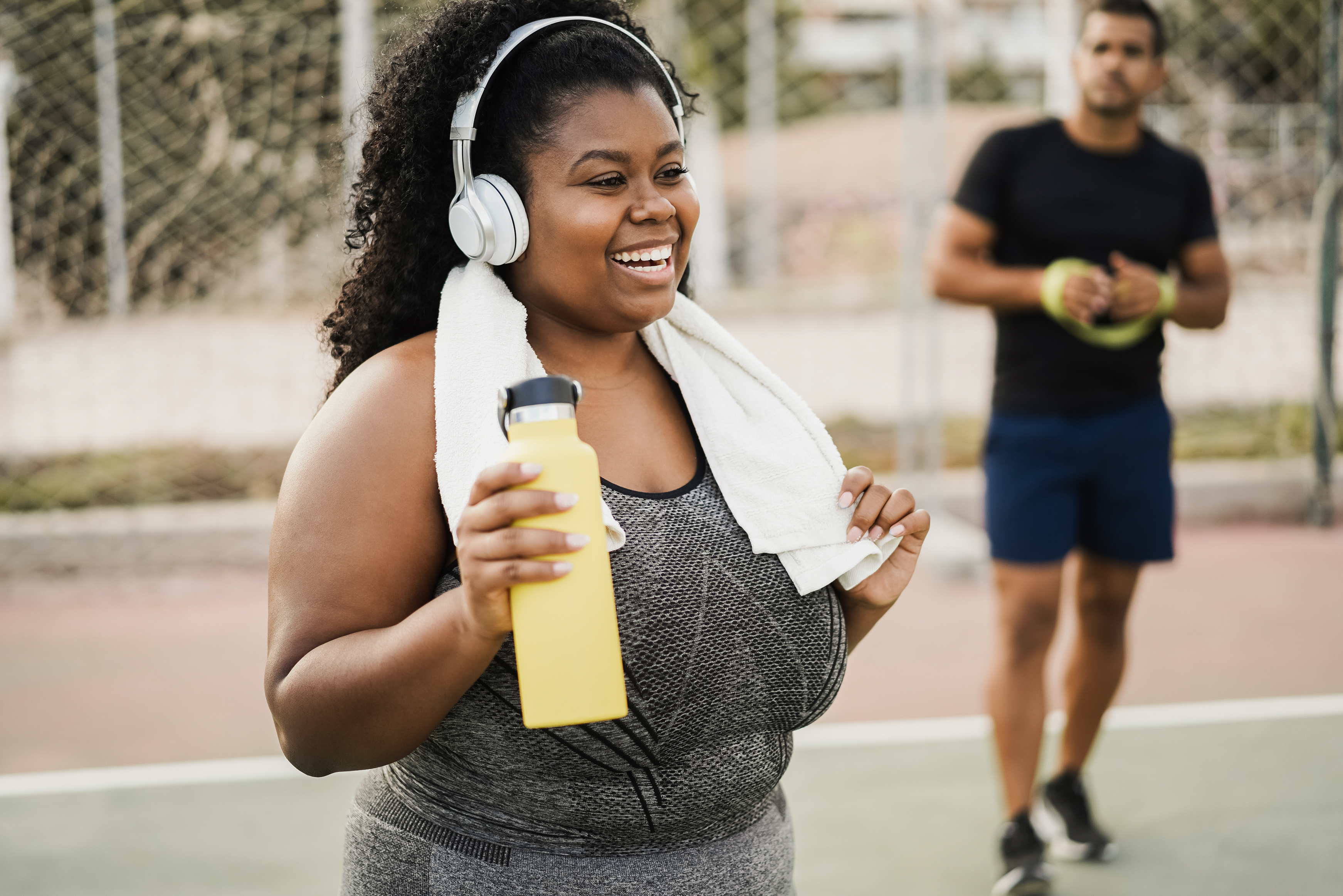 A smiling woman wearing headphones and holding a water bottle wipes her hand on a towel draped over her neck. She is standing on an outdoor court, with a man holding an exercise band standing behind her.
