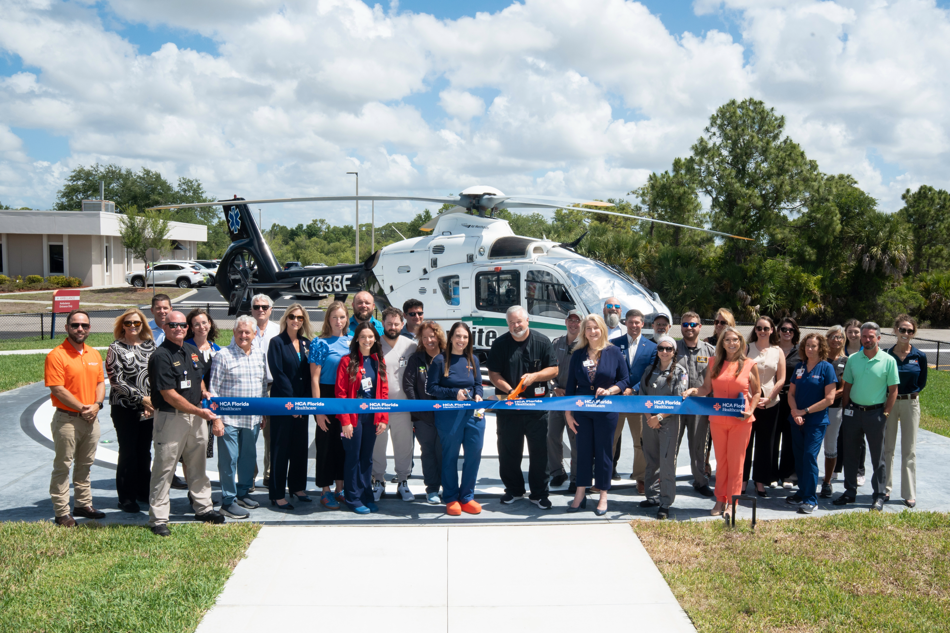 Hospital leaders, emergency care teams, EMS crews and community partners gather for a ribbon-cutting ceremony celebrating the opening of the new helipad at HCA Florida Englewood Hospital. A medical helicopter is positioned behind the group during the outdoor event recognizing expanded emergency transport capabilities.