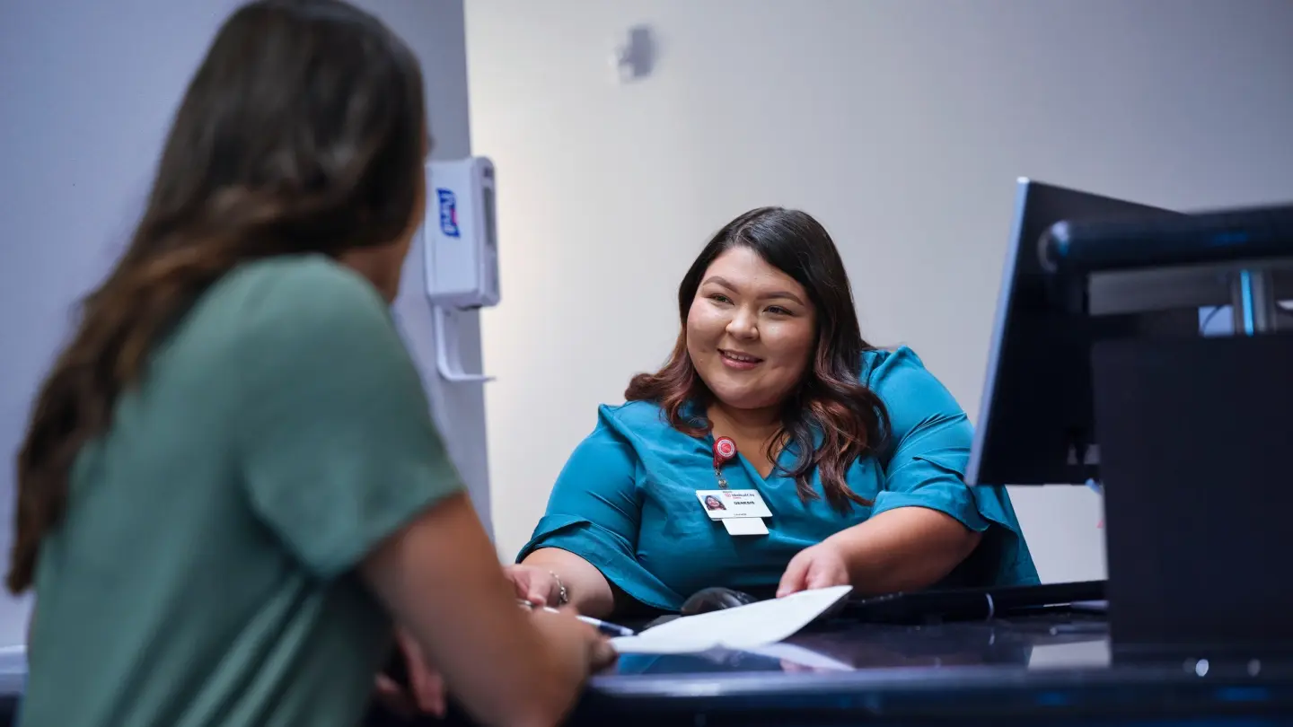 A female hospital staff member sitting at a desk reviewing paperwork with a female patient. 
