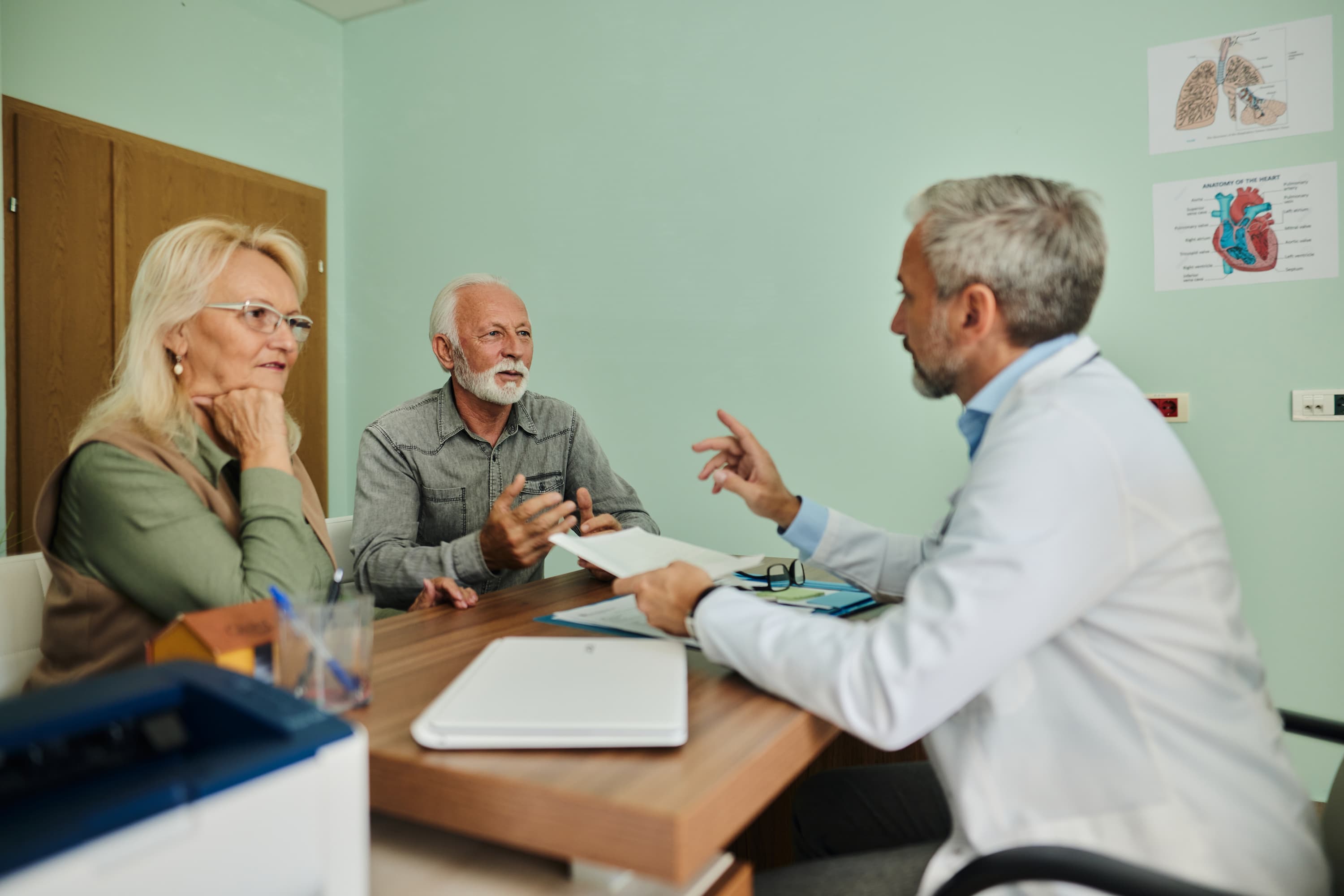 A doctor in a white coat talks with an older couple seated across a desk in a medical exam room.