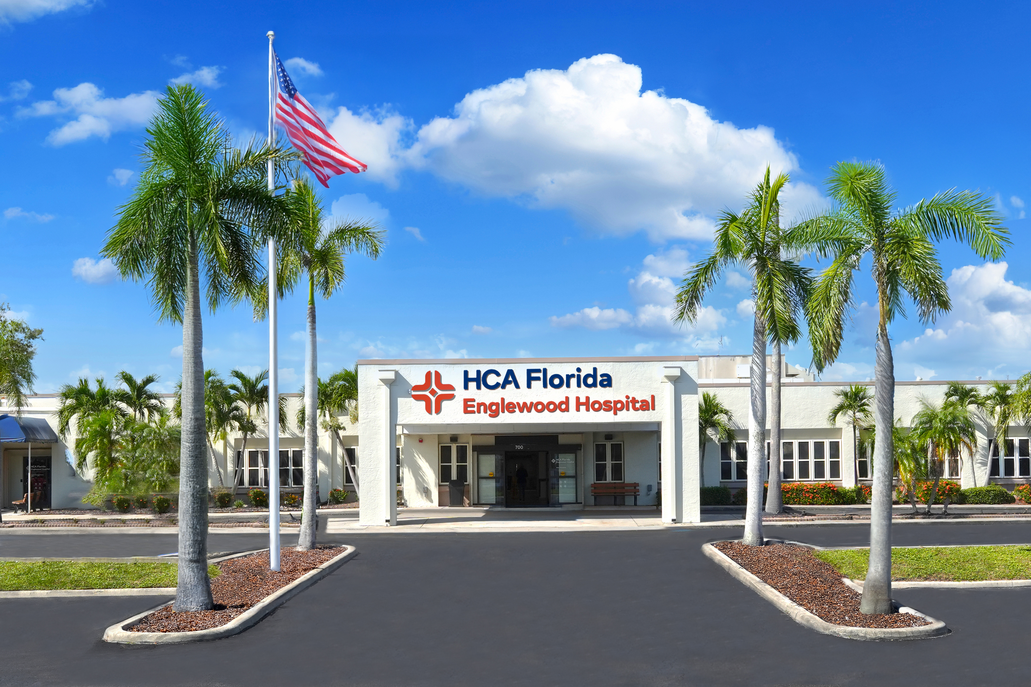 Main entrance of HCA Florida Englewood Hospital with a white facade, red and blue logo signage, American flag on a pole, and palm trees lining the driveway under a bright blue sky with scattered clouds.