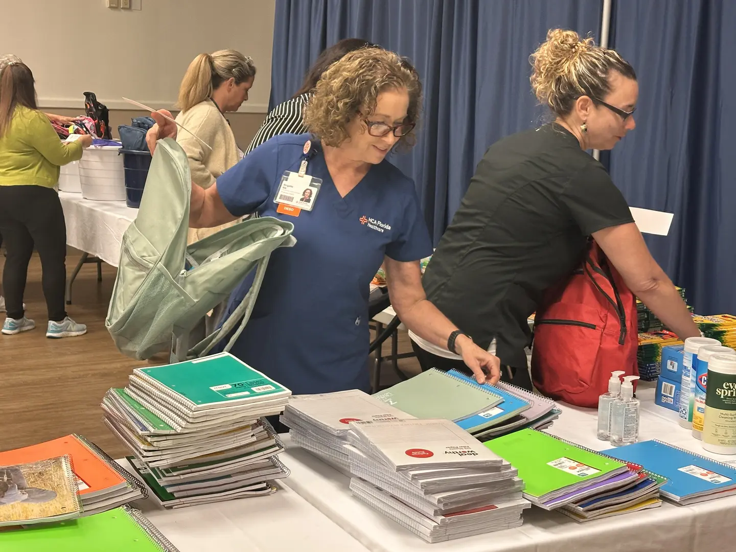 A group of people in a room organizing school supplies on tables. One person in a blue shirt holds a light green backpack, while another in a black shirt reaches for items. The tables are filled with notebooks, folders, hand sanitizer, and disinfectant spray. More individuals are seen in the background near another supply table.