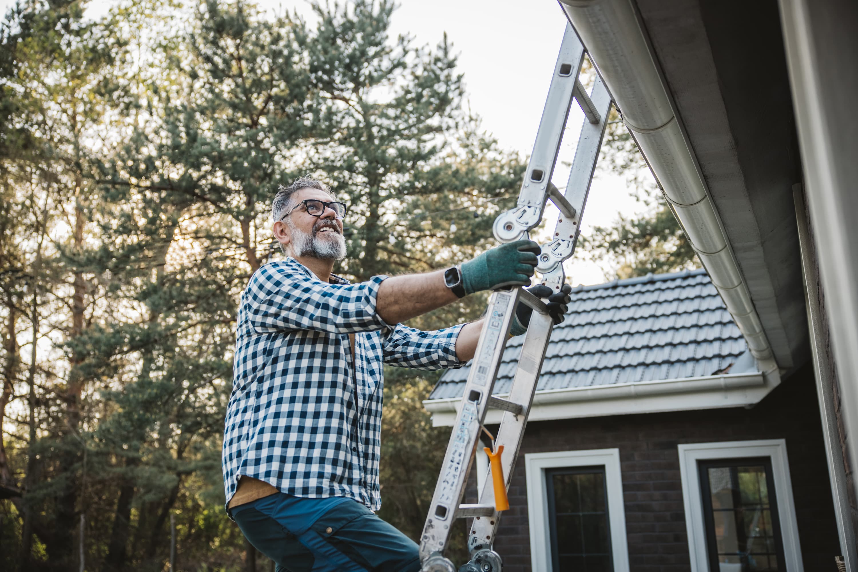 A smiling gray-haired man in glasses and work gloves adjusts an extension ladder beside a house gutter, with trees and another house in the background.