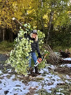 Dana DeBernardi carrying a large tree branch over her shoulder, with a chainsaw in her other hand.