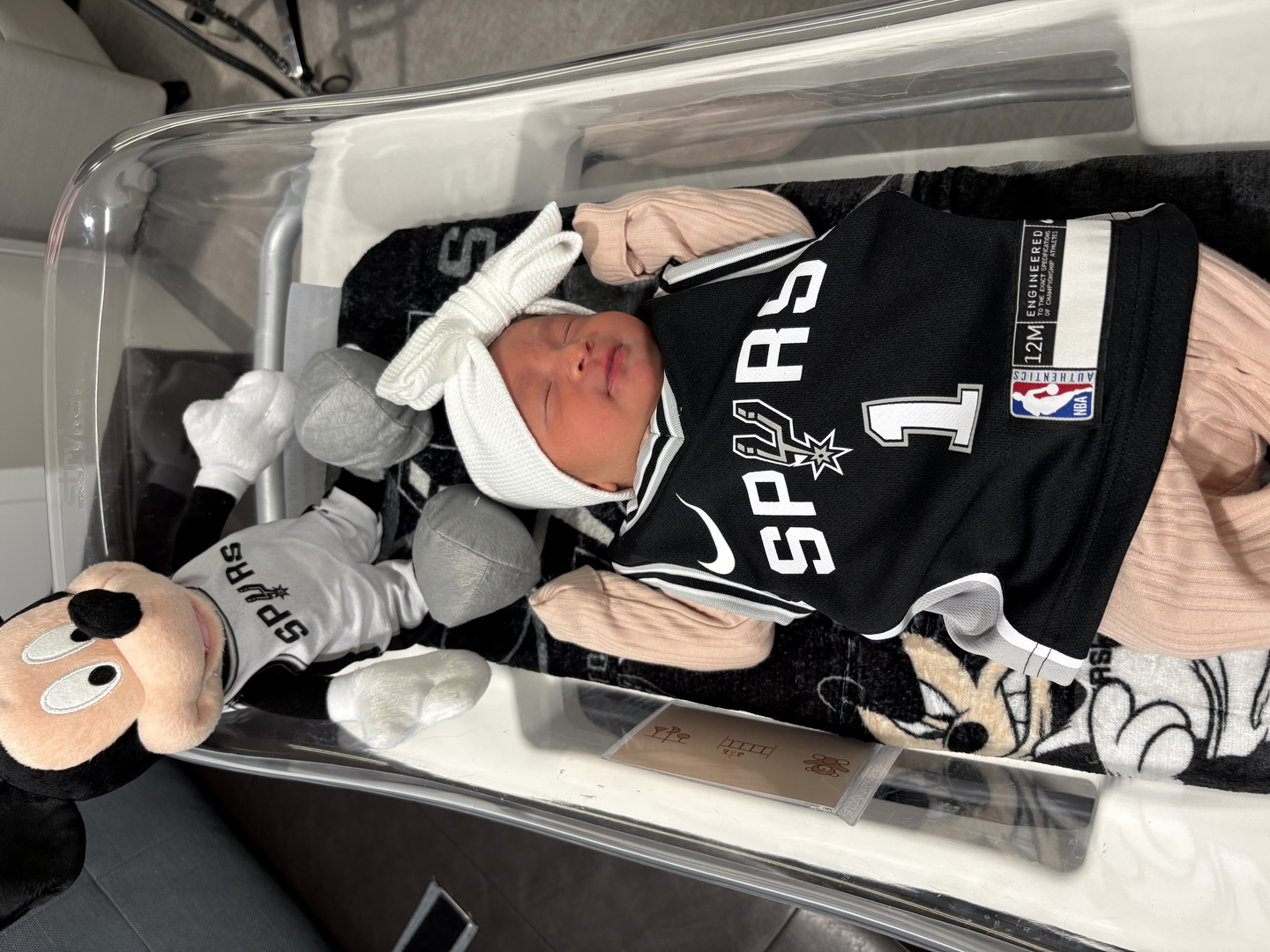 Newborn baby in hospital crib, wearing a spurs jersey and a spurs plush toy.