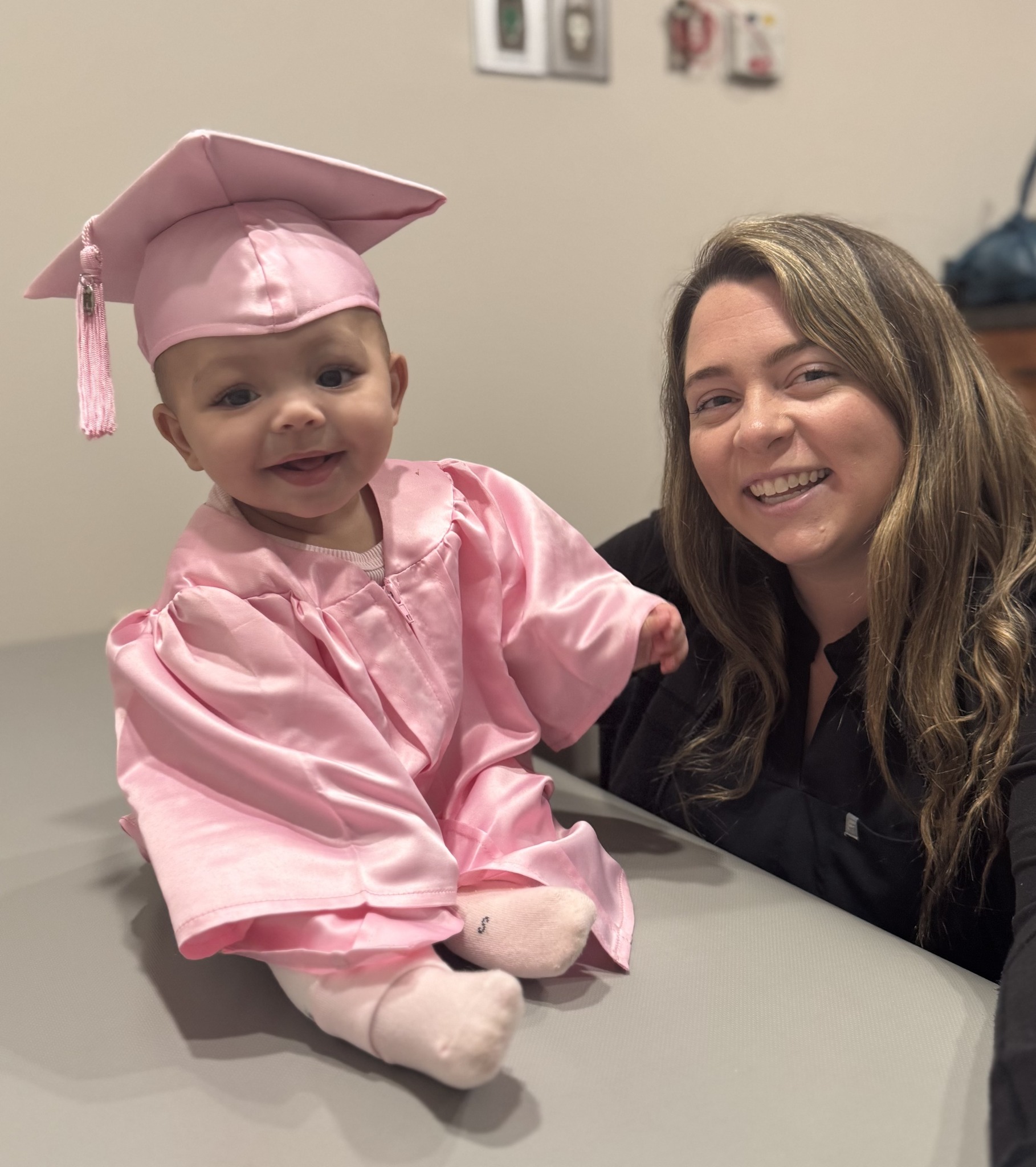 A baby in a tiny pink graduation gown and cap with with her physical therapist smiling beside her