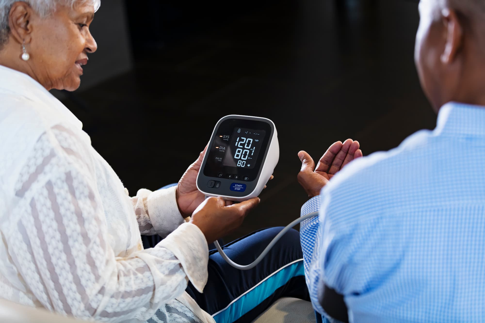 Older adult using a home blood pressure monitor on another person, with the digital screen displaying a reading.