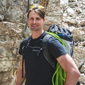 Matt Berry smiles in front of a rock wall, with a backpack and neon green rope on his back.