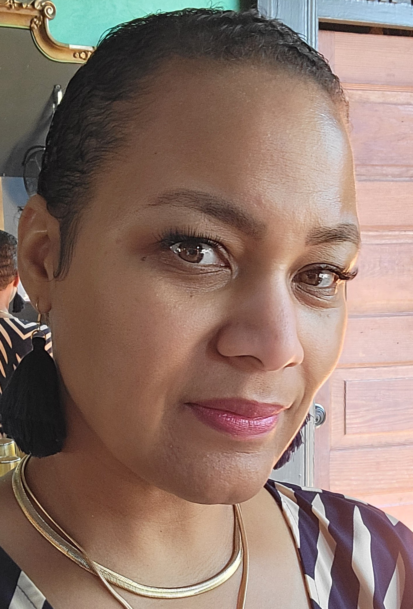 Close-up portrait of a Tinesha indoors wearing a patterned V-neck top, layered gold necklaces, and earrings, with her hair pulled back and soft natural makeup, looking slightly to the side.