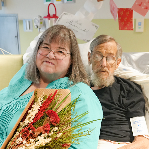 Marlene sits with William in hospital bed, holding bouquet of flowers.