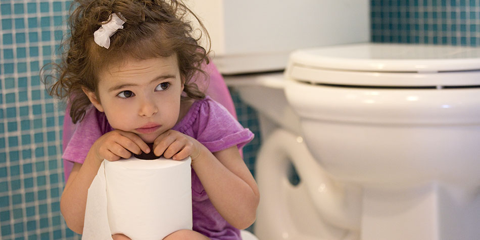 A little girl sits on the toilet, looking sad while holding a roll of toilet paper.
