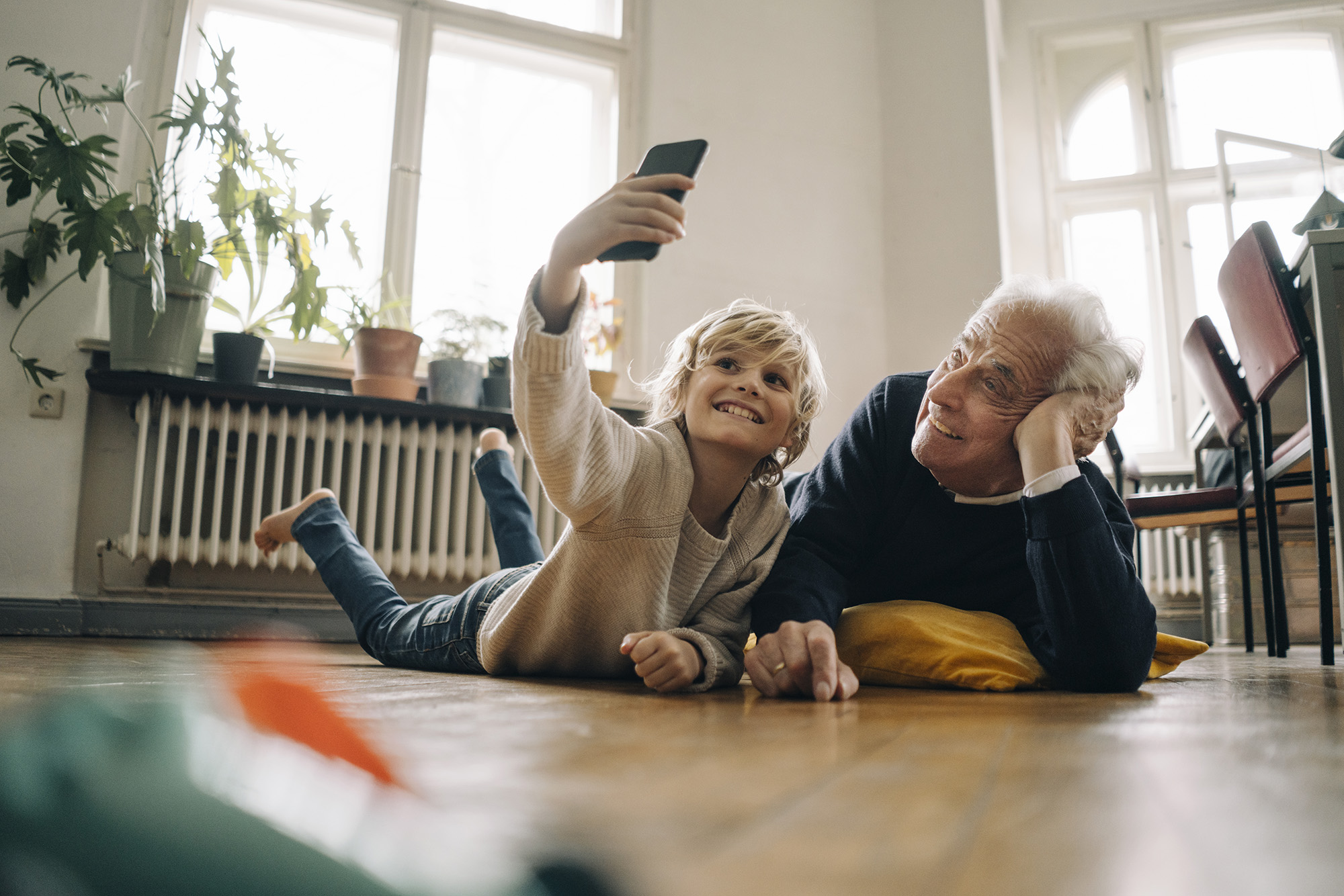 An older man and a child lying on the floor at home, taking a selfie with a smartphone.