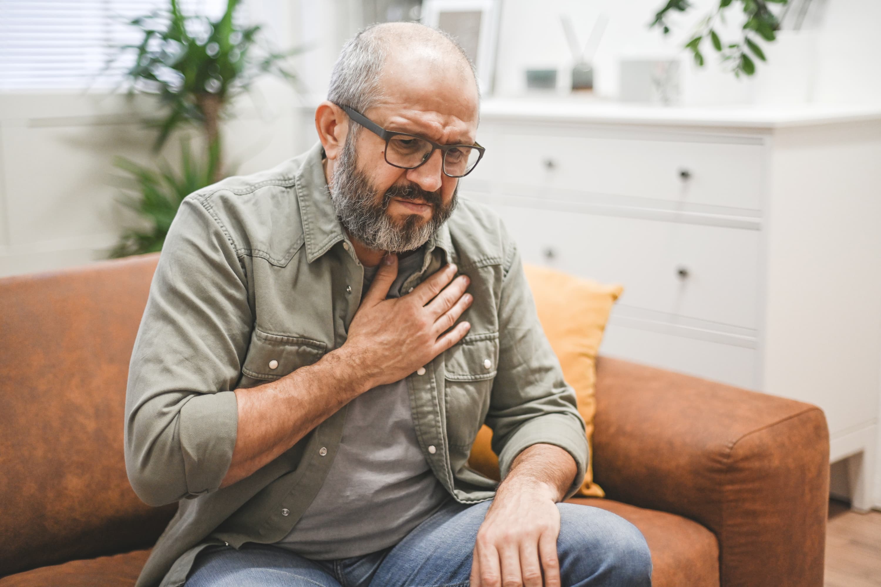 Older man sitting on a couch, holding his chest in pain and appearing distressed.