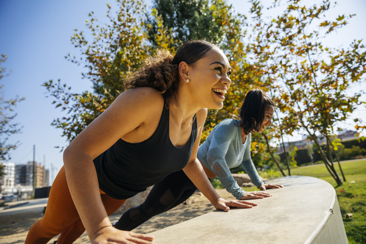 Two cheerful women completing push-ups on a retaining wall at a park.