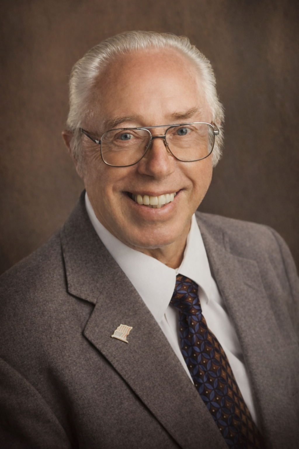 Professional headshot for Woody Andersen. He is wearing rimmed glasses, a grey/brown suit jacket with a tie, a white dress shirt underneath, and an American flag pin on his lapel.