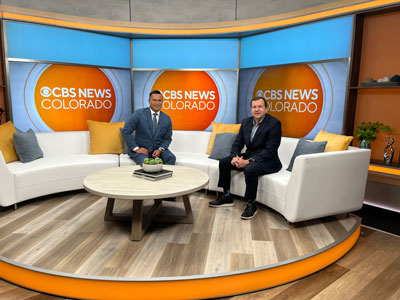 Two men sitting on a couch at a news station smiling at the camera.