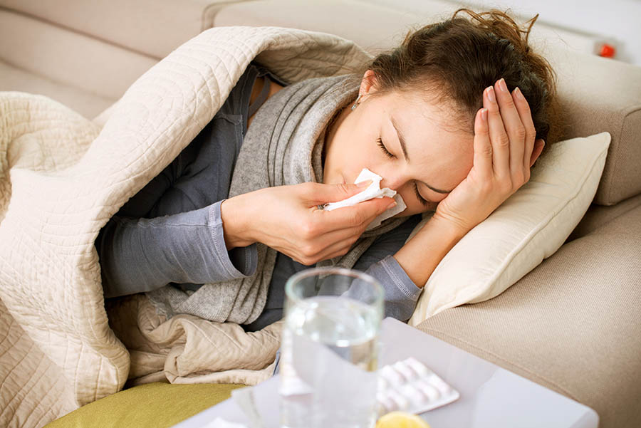A woman lies on the couch with a blanket, while blowing her nose.