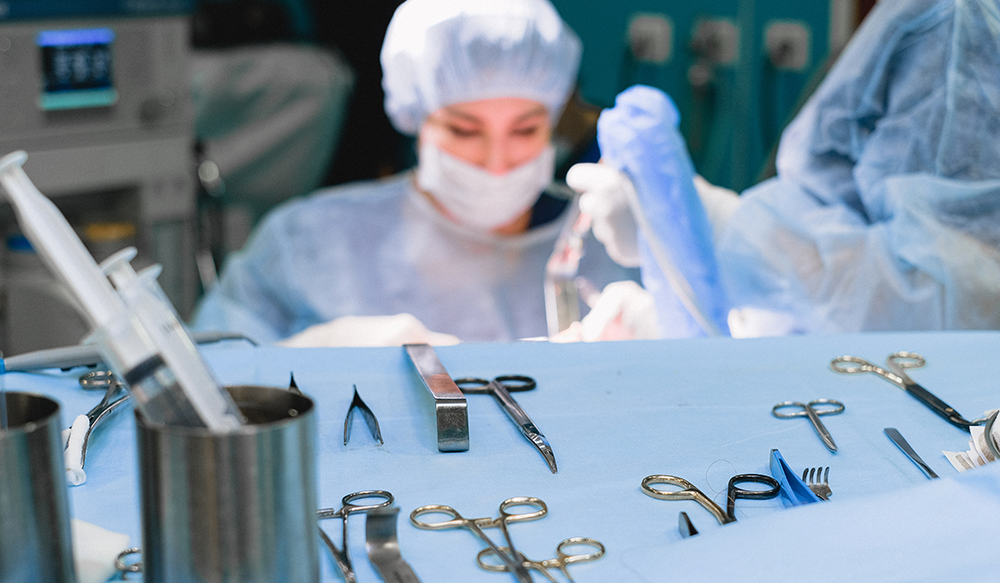 Operating instruments sit on a table, while surgeons wearing surgical scrubs operate in the background.