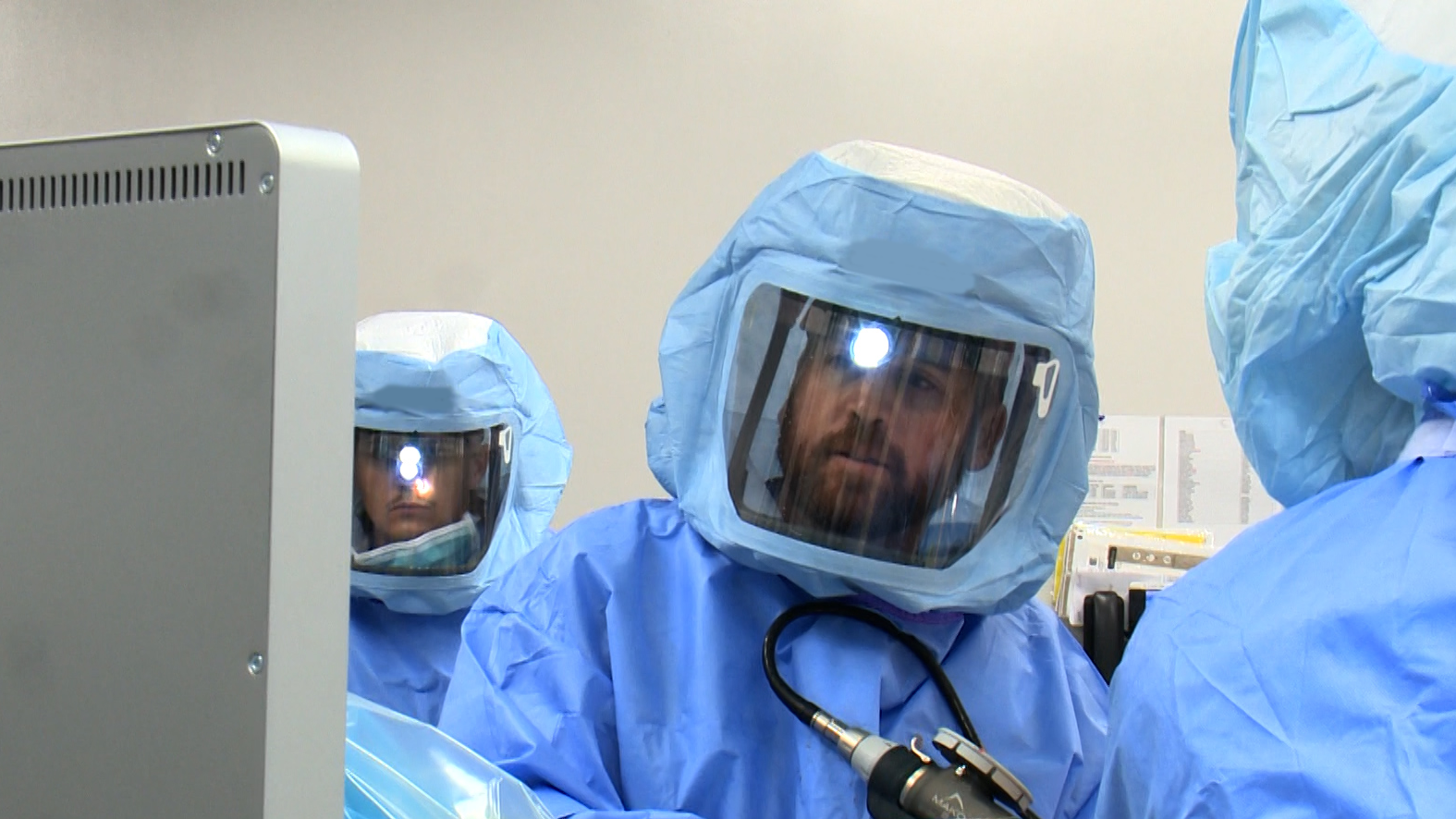 Surgeon is in an operating room with two other people and the patient. The surgeon's face is the only face showing. He is performing a surgery using the new robotic surgical equipment in his hand.