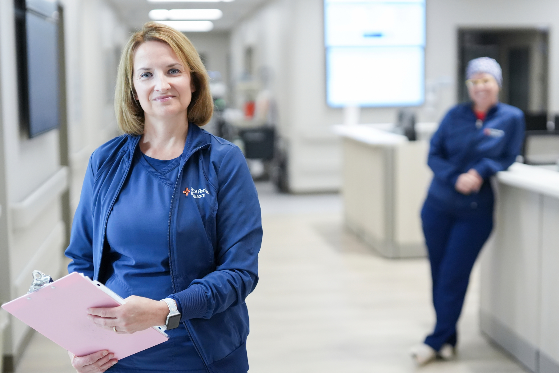 A healthcare professional wearing blue HCA Florida Healthcare scrubs stands in a hospital hallway holding a clipboard, with another colleague visible in the background near a nurses station.