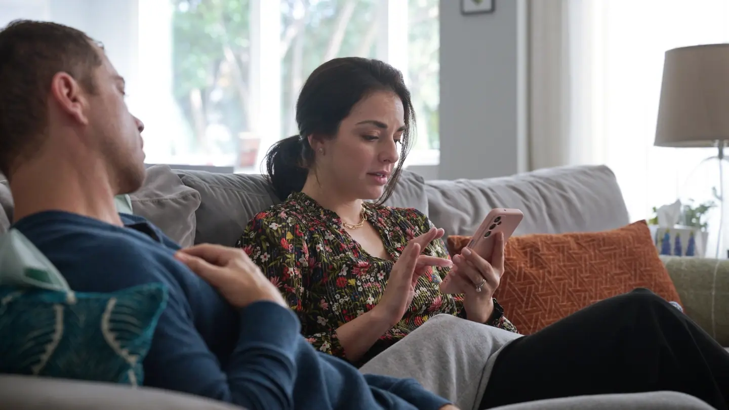 A couple sitting on the couch. The male is laying with his legs over the woman while the woman is scrolling on her phone.
