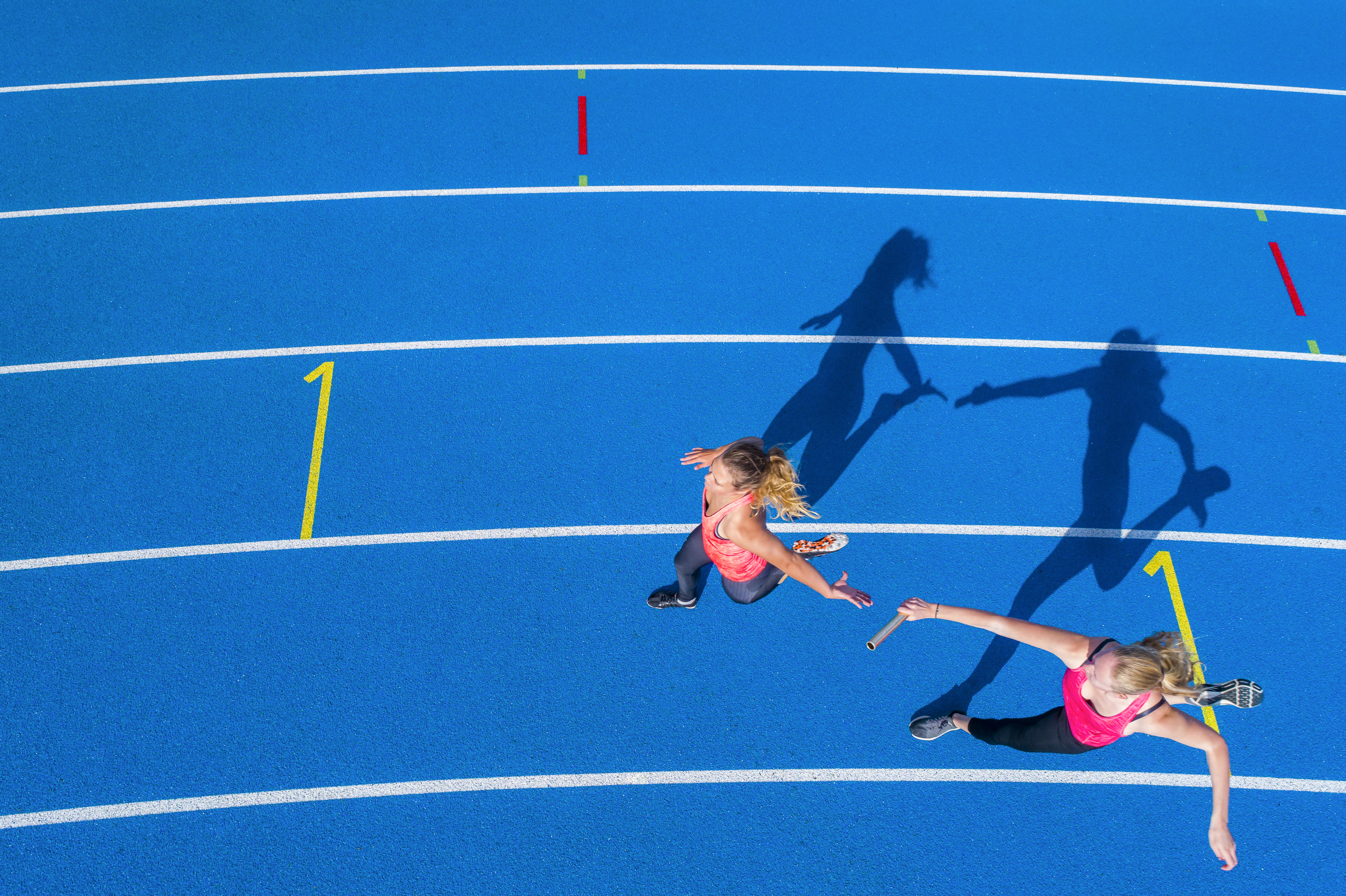 Top view of two female runners passing the baton on a blue running track.