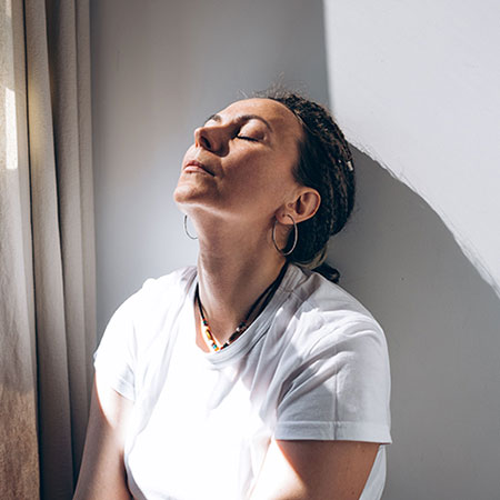 A woman leaning back against a wall. She has her eyes closed with sunshine from the window casting over her face.