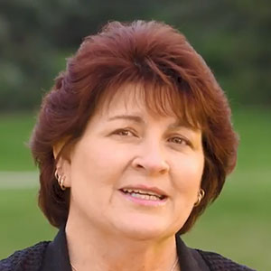 Gayle smiles while standing outside in a park, wearing a black cardigan.