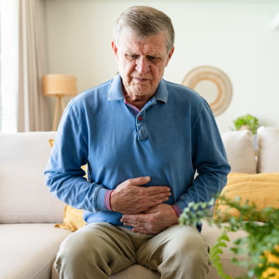 An older adult seated on a sofa in a bright living room holds both hands over their lower abdomen with a pained expression, suggesting stomach discomfort. A floor lamp, decorative wall piece, cushions, and houseplants are visible in the background.