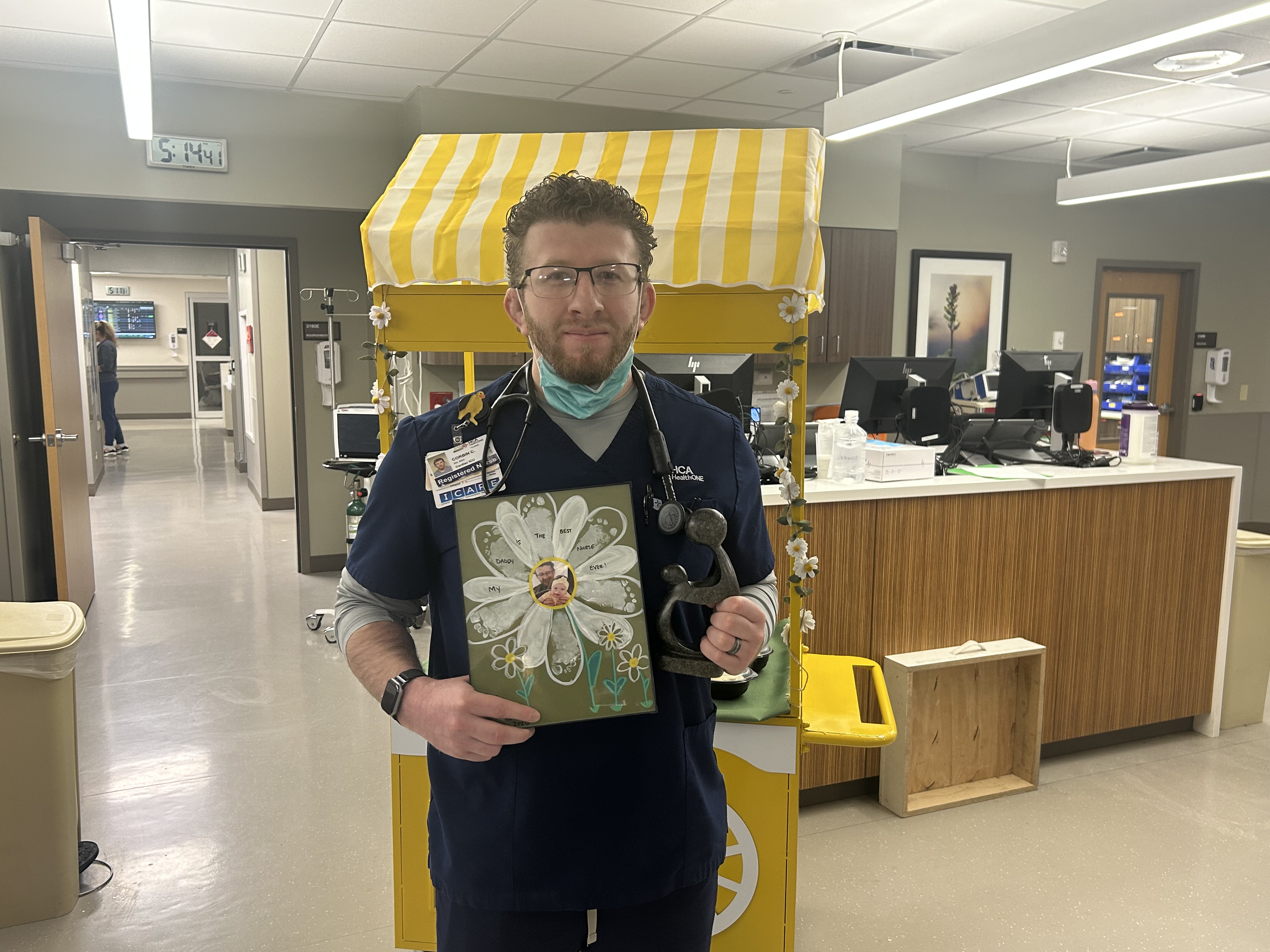 Corbin Chavez in scrubs standing in a hospital unit, holding a decorative art piece with a large white flower.
