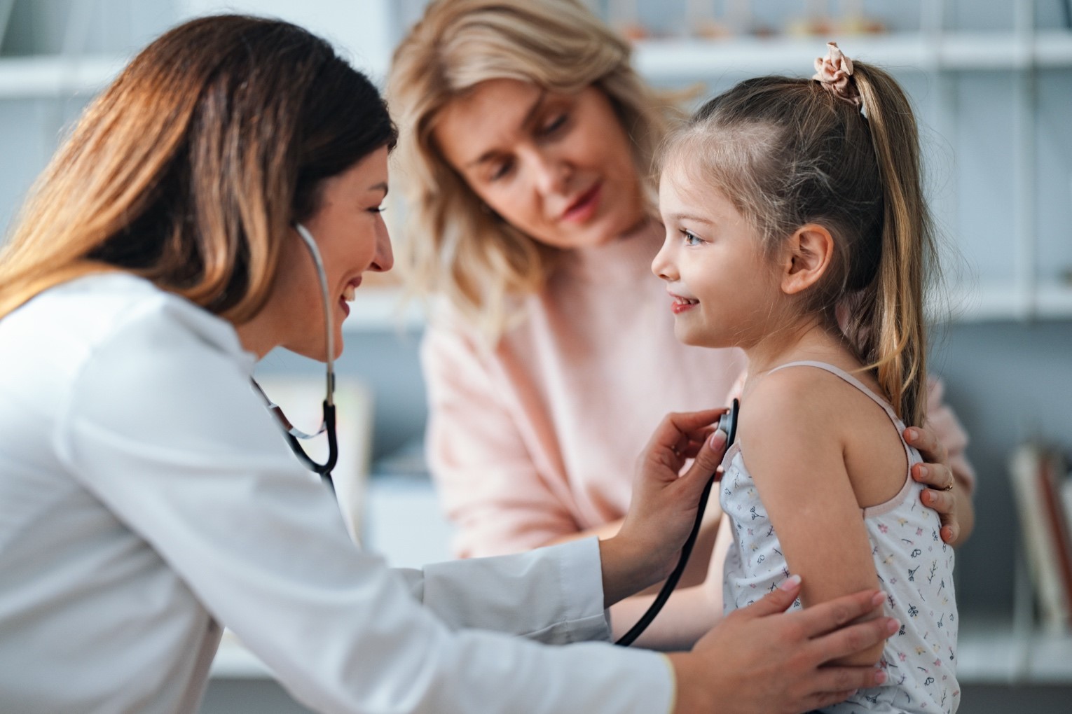 A pediatrician examining a young girl with a stethoscope in a medical office. The girls mother is standing beside her with her arm on the girl's back.