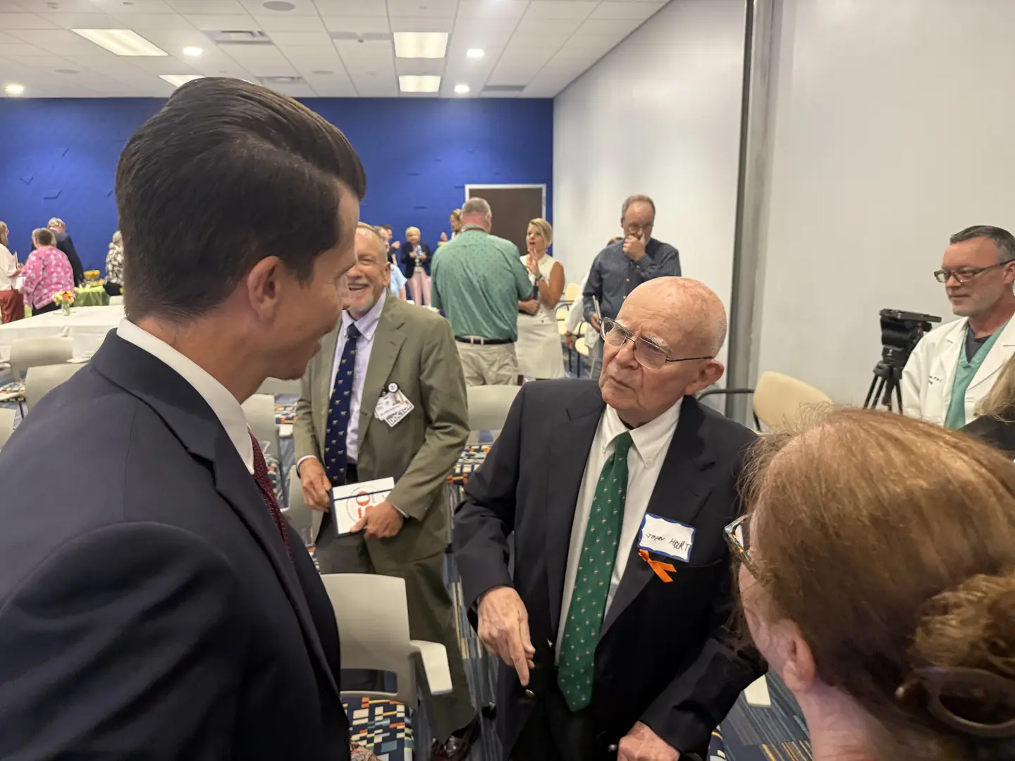 On the left is the back of a tall man with dark hair. He is facing a man who is talking to him. The man talking is wearing a dark suit and tie. The two men are surrounded by other people in the room.