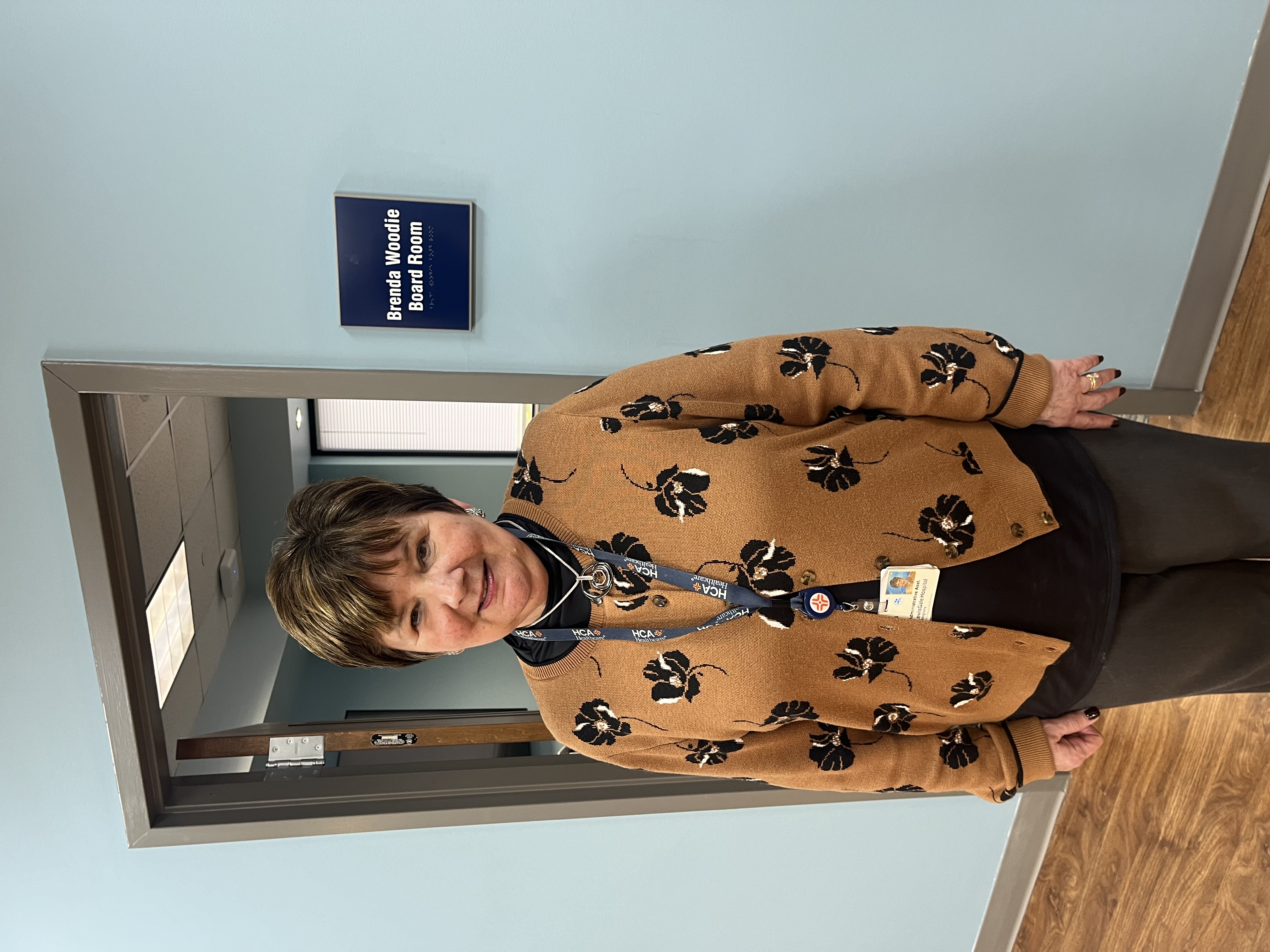 Brenda Woodie smiles while standing proudly in front of a plaque proclaiming a meeting room the Brenda Woodie Board Room.
