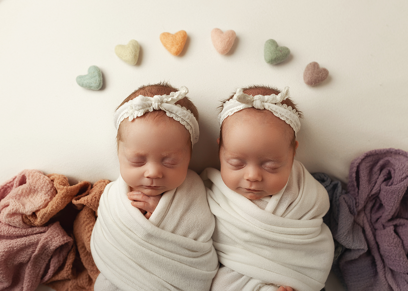 Nygaard twins professional portrait, swaddled in blanket with a rainbow of hearts above their heads.