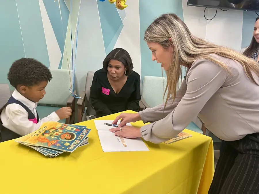 Young boy Payton seated at table with books surrounded by female hospital staffer and his mother.