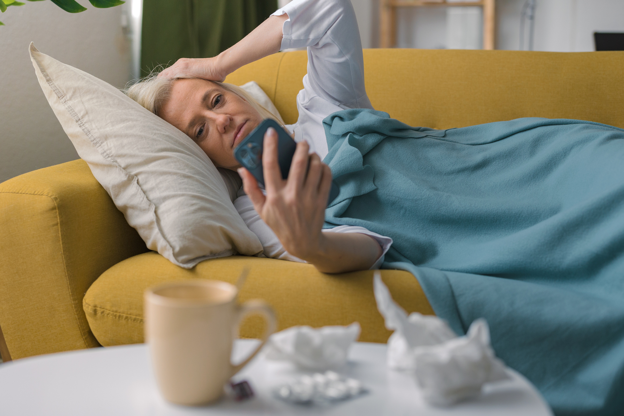 Ill woman lying on a sofa, looking at her smartphone.